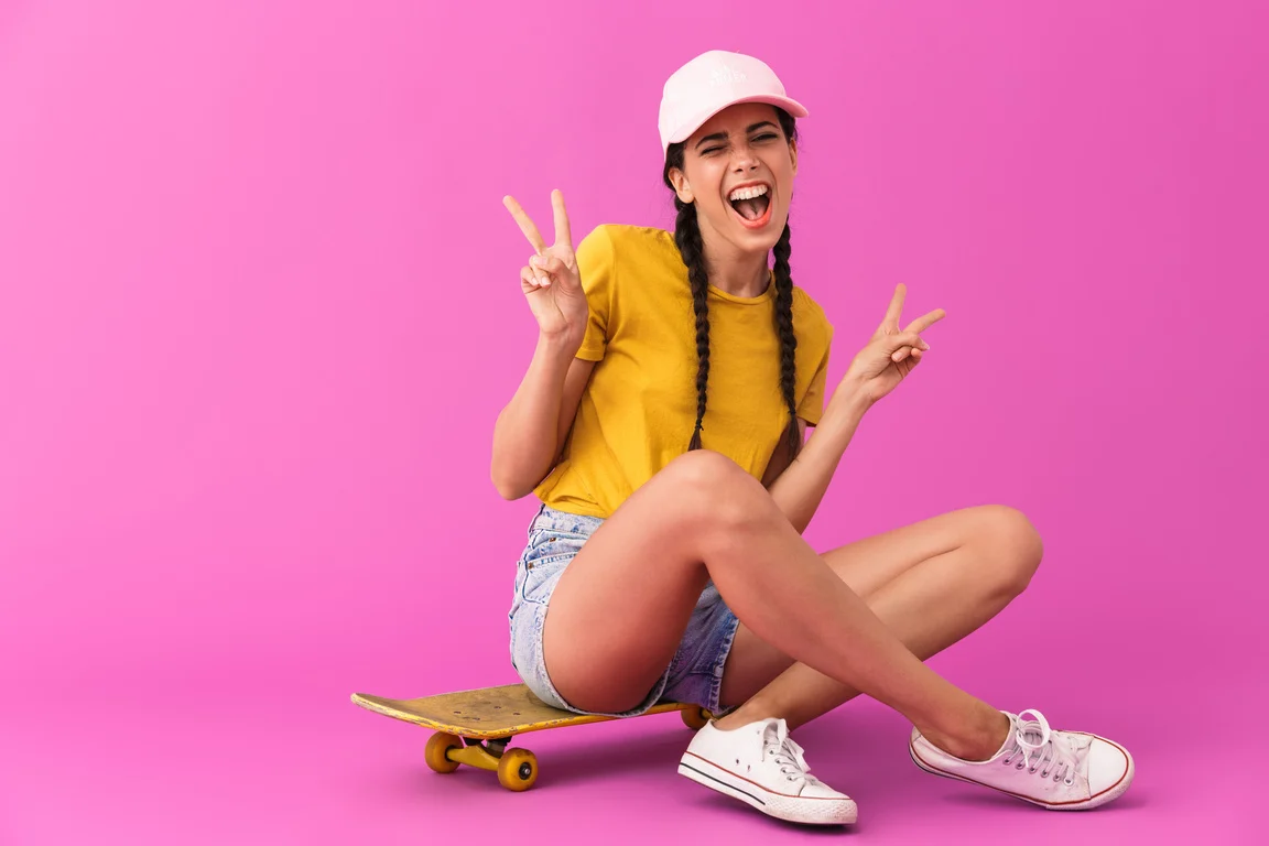 Image of Smiling Happy Woman Wearing Casual Clothes Showing Peace Sign and Sitting on Skateboard